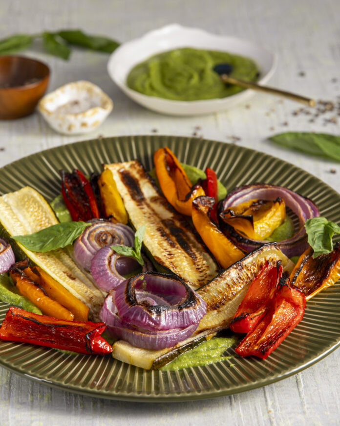 A green plate with red, yellow, green, orange, and purple grilled vegetables garnished with fresh basil and a small white bowl of pesto on the side.