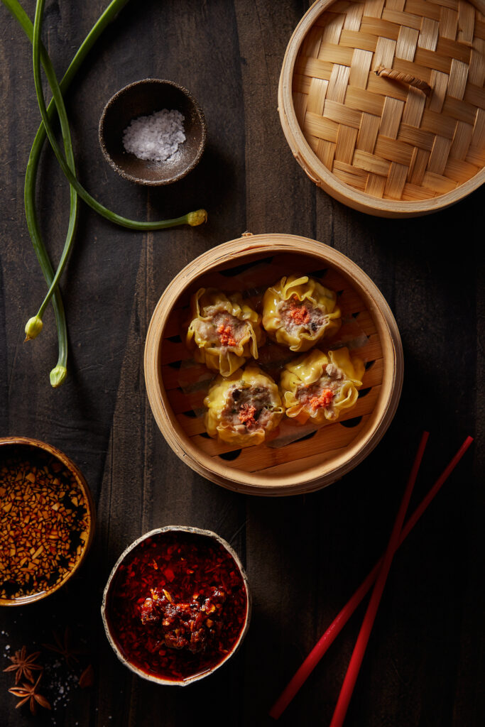 Four dumplings sit in a round wooden bowl, two bowls of sauce sit off to the left.