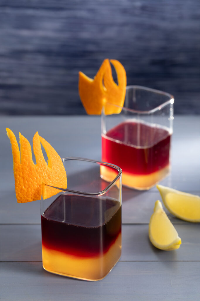 Two layered cocktails, orange on the bottom and red on the top, in clear square glasses with citrus flames as garnish and 2 lemon wedges to the right of the glasses.