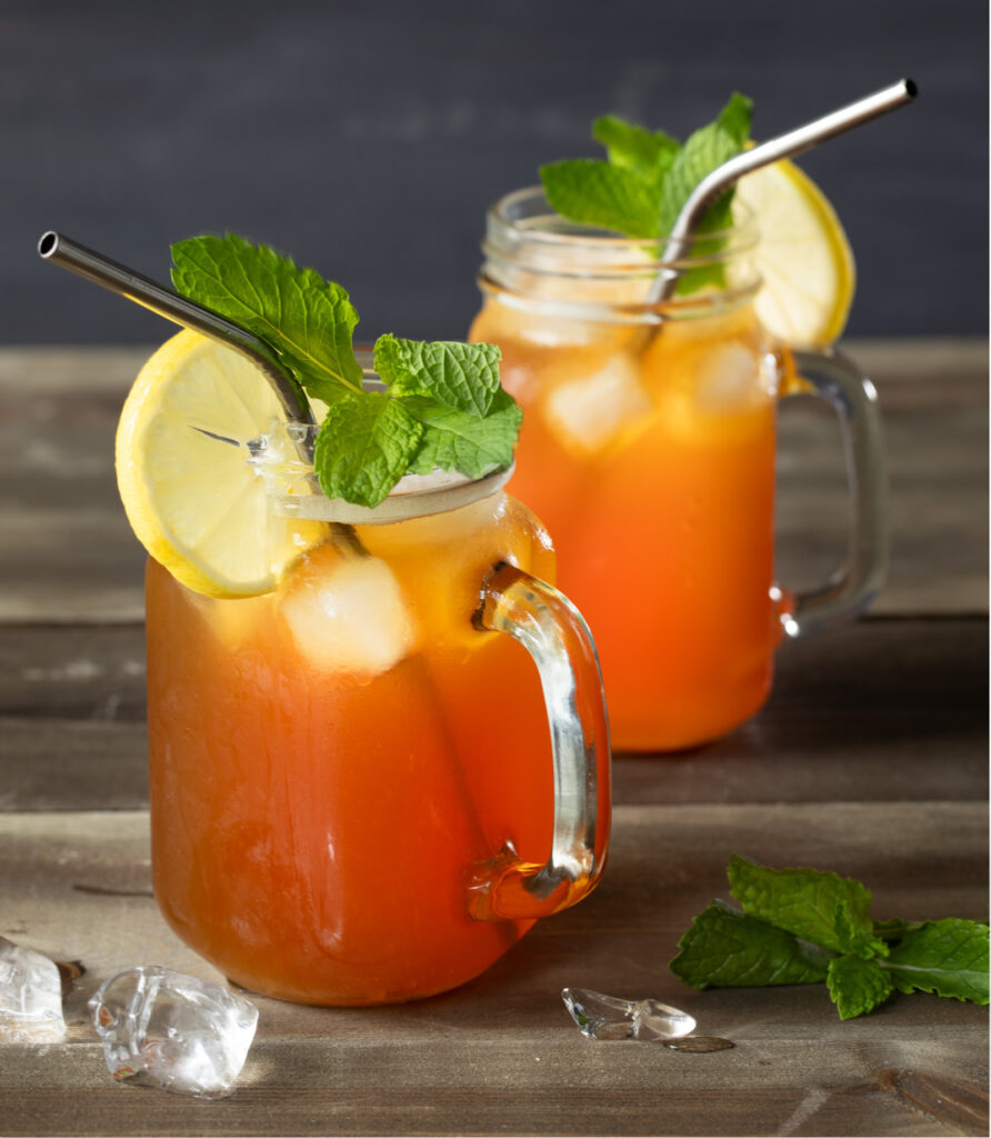 Two mason jar mugs with what looks like ice tea garnished with lemon and mint on a wooden surface with melting ice. cubes