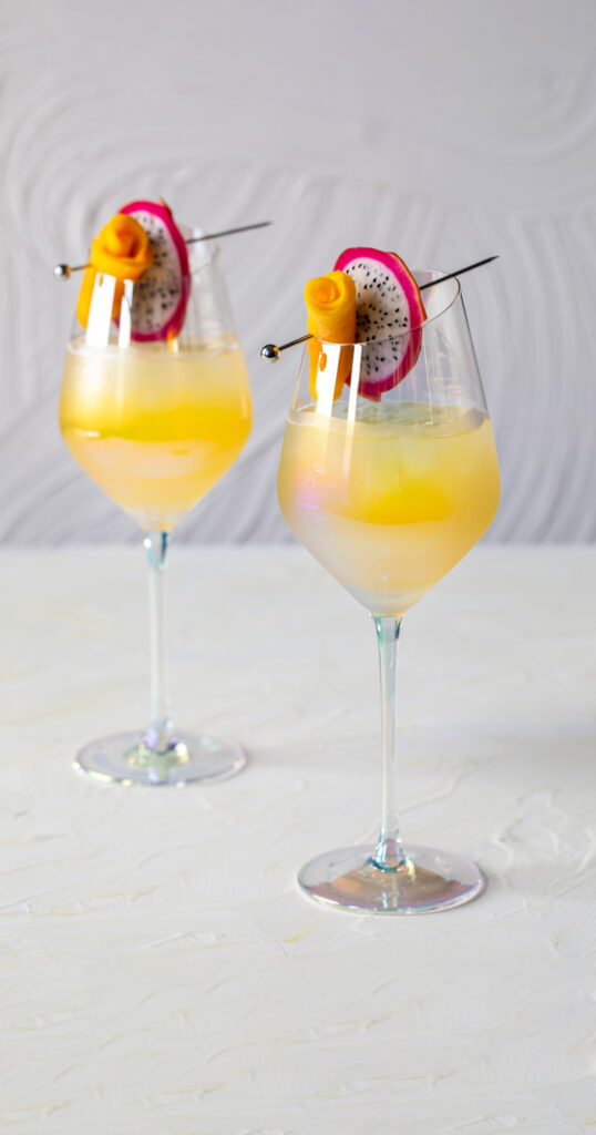 Two identical cocktails in iridescent wine glasses garnished with dragon fruit pinwheels and mango rolls made to look like roses.