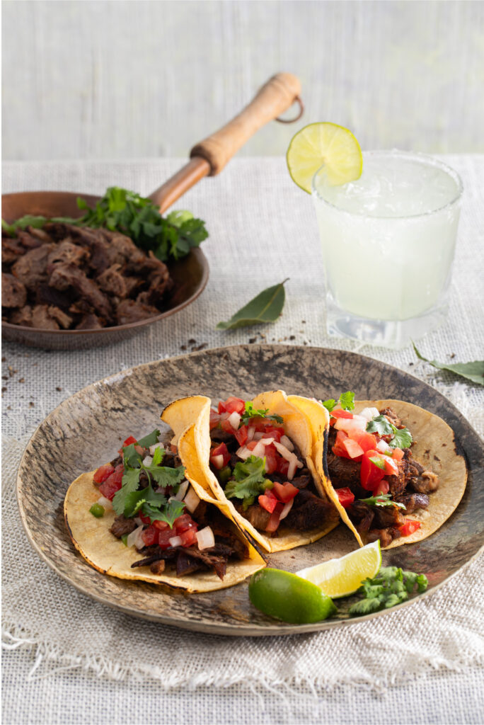 Three Tacos de Lengua sit on a plate. Red tomatoes, onion, and cilantro are seen on top. Tacos de Lengua Recipe