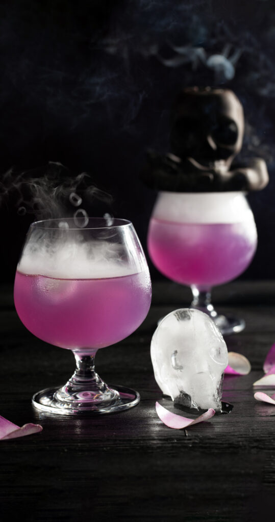 Two light purple cocktails with smoke and skull ice cubes on a black background