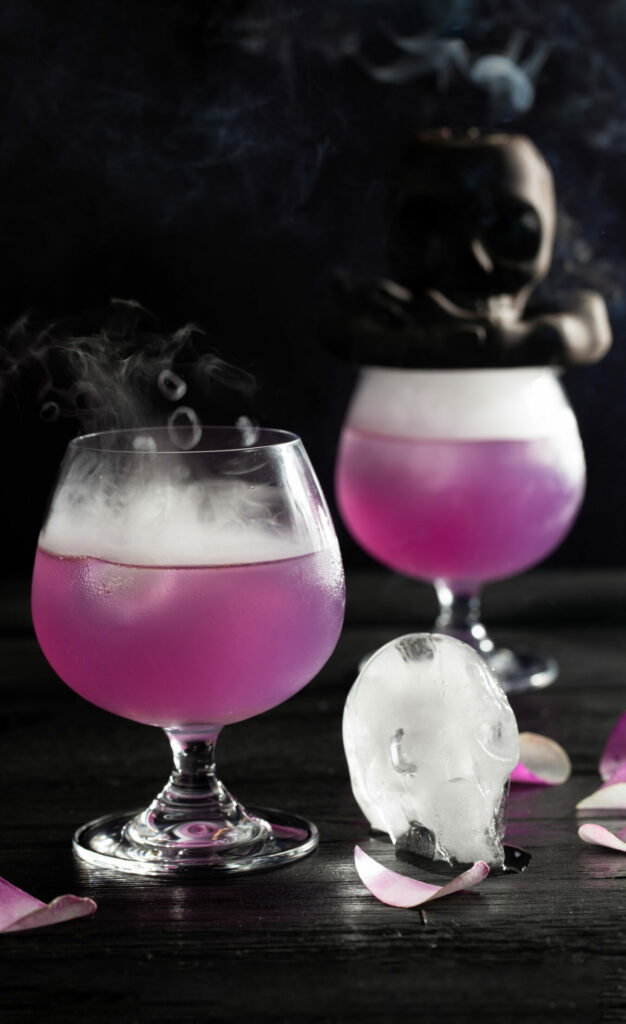 Two light purple cocktails with smoke and skull ice cubes on a black background