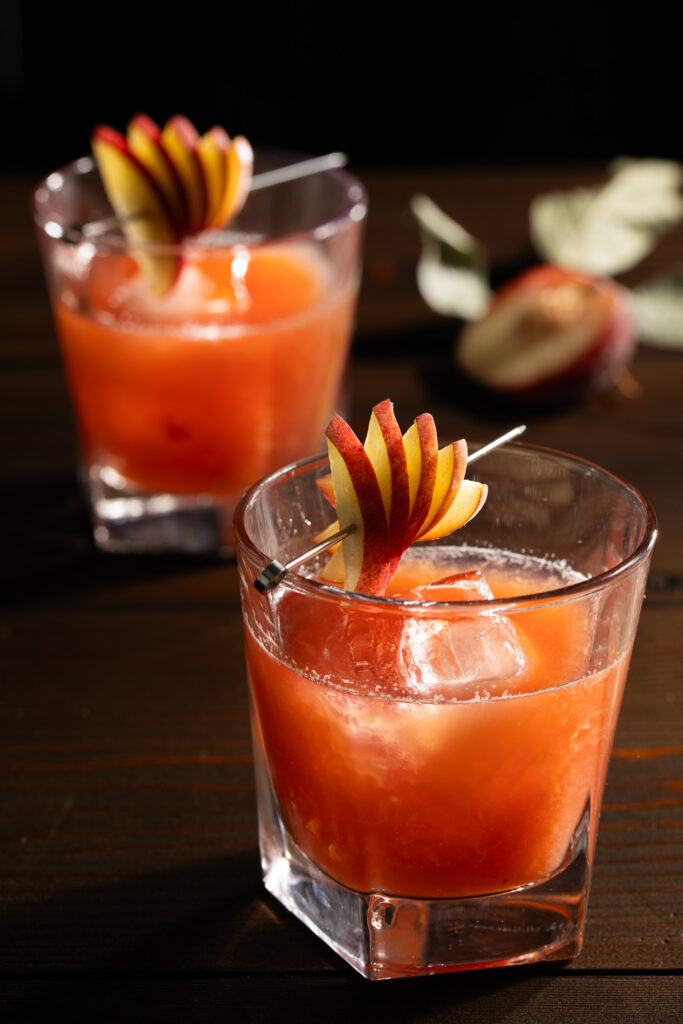Two orange colored cocktails in rocks glasses with peach fan garnish and a dark background