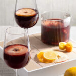 Two deep red sangria cocktails sit in wine glasses and garnished with lemon wedges. Sangria Recipe