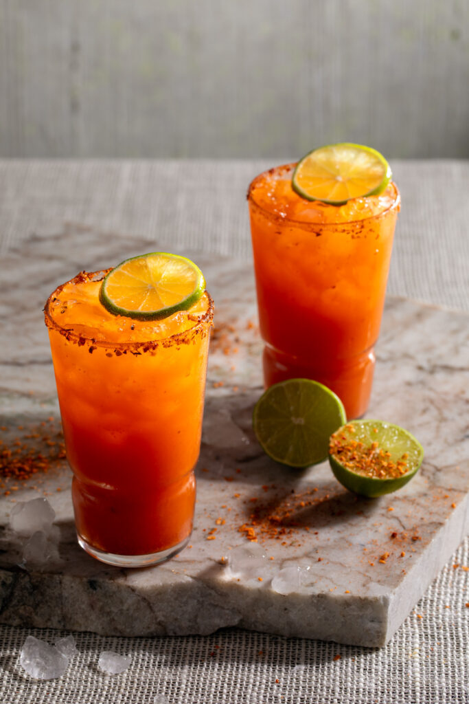 Two Michelada cocktails orange red in color sit in tall glasses, garnished with lime and Tajín. Michelada recipe