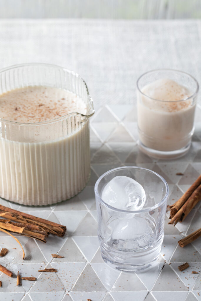 Horchata, a frothy beverage light brown in color, sits in a pitcher next to two glasses, one filled with Horchata, one with just ice. Horchata Recipe