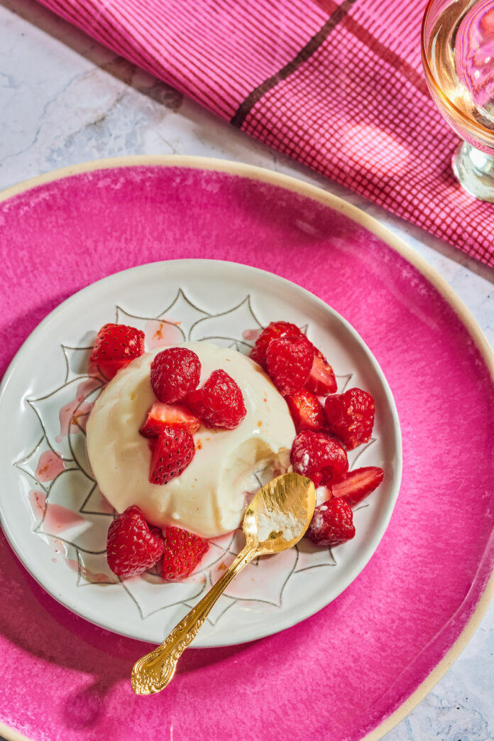 Rose Panna Cotta with topped with strawberries on a white plate, sitting on a larger pink plate. a gold spoon sits to the right side of the rose dessert.