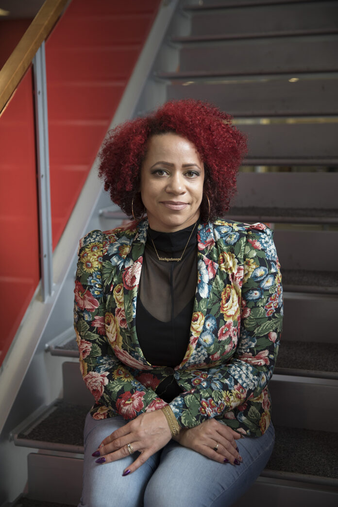 Nicole Hannah-Jones, a speaker in AWAACC's TRUTHSayers series sits on a set of stairs with a colorful printed blazer and red, curly hair.