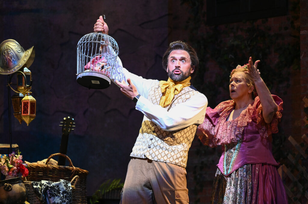 a man holds up a bird cage with a weird expression on his face. a woman stands behind him with his hands up