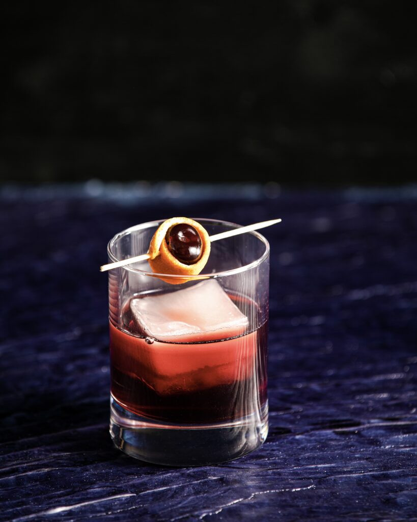 A dark blue and black background with a red, Cherry Tart Negroni cocktail with a square ice cube, garnished with a cherry wrapped in orange peel on a pick.