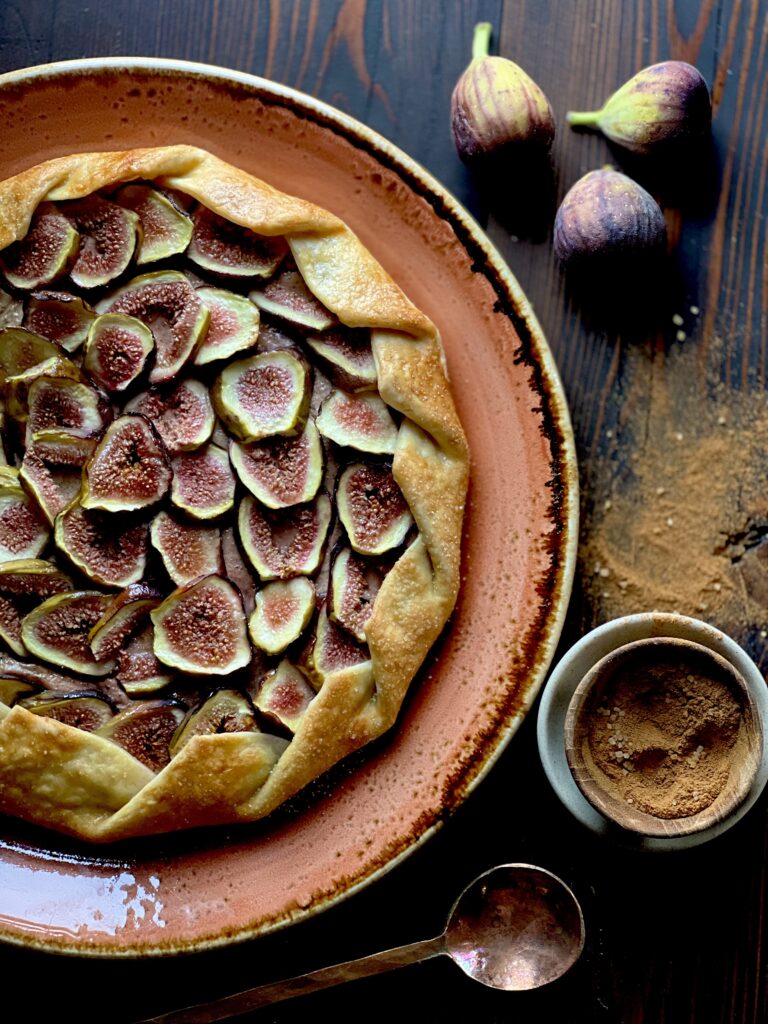 A galette with sliced figs on a terra cotta colored plate with figs and spice on a dark surface