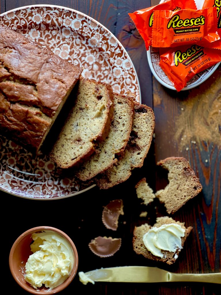 A partially sliced loaf of peanut butter cup banana bread on a brown and white floral plate with a small bowl of whipped butter and a little dish of wrapped Reese's peanut butter cups.