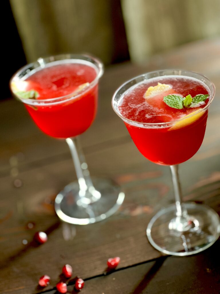 Two cocktails, bright ref in color sit in wine glasses and are garnished with lemon round, pomegranate arils, and mint leaves
