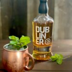 A copper mug with a cocktail garnished with fresh mint next to a bottle of Dubliner Steelers Select Irish Whiskey on a brown wood surface.