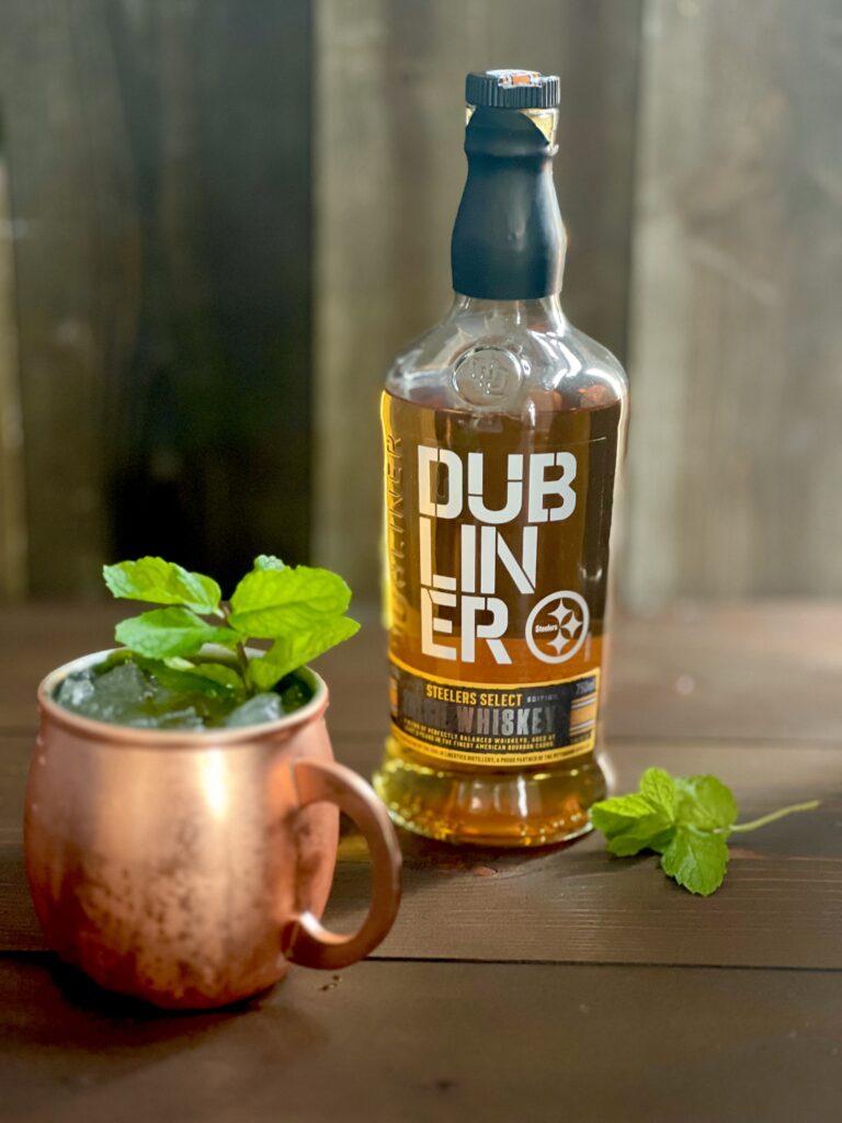 A copper mug with a cocktail garnished with fresh mint next to a bottle of Dubliner Steelers Select Irish Whiskey on a brown wood surface.