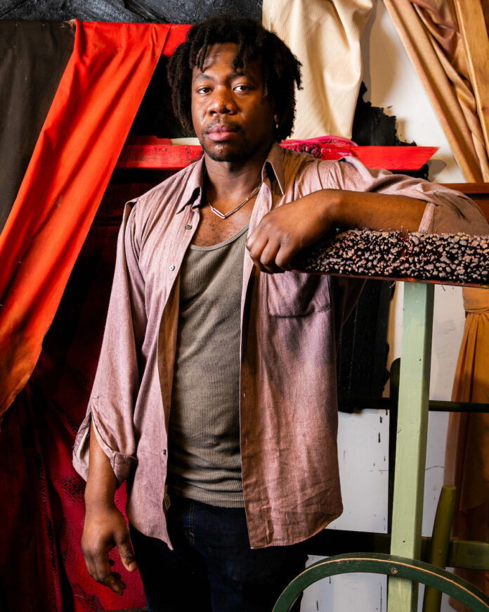 a black man with short dreads leans against something that looks like a cat scratcher in his studio. artist Steve Alexis
