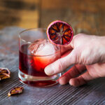 A hand comes from the right of the frame and holds a Blood Moon Negroni Cocktail, red in color. Garnished with a dried blood orange wheel. Blood Moon Negroni Cocktail Recipe
