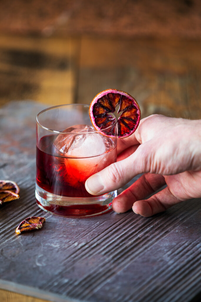 A hand comes from the right of the frame and holds a Blood Moon Negroni Cocktail, red in color. Garnished with a dried blood orange wheel. Blood Moon Negroni Cocktail Recipe
