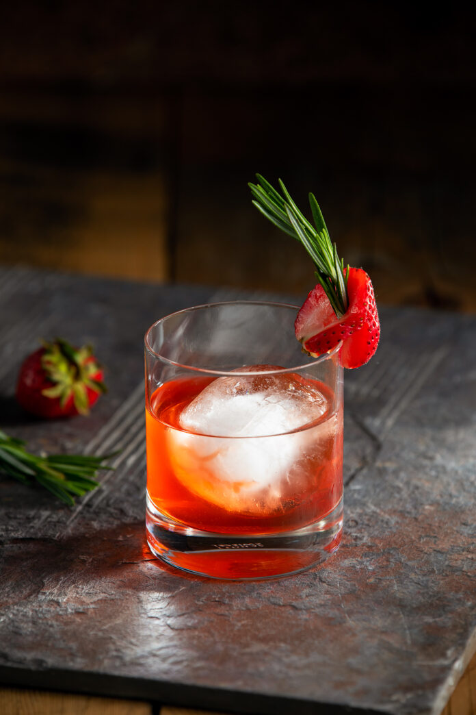 A cocktail orange in color sits in a rocks glass, garnished by a strawberry. Punch Drunk Negroni Cocktail Recipe
