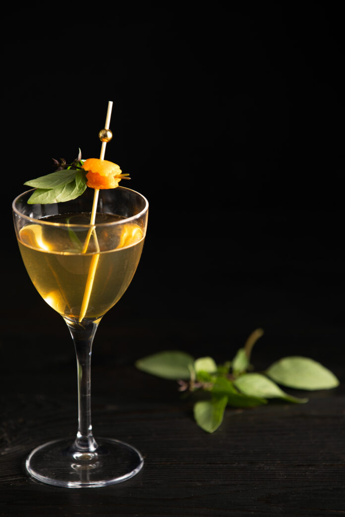 A pale yellow cocktail in a wine glass garnished with basil