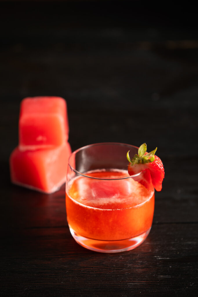 A red cocktail in a rocks glass sits next to two frozen red fruit cubes. The Beauty School Dropout Negroni Cocktail Recipe