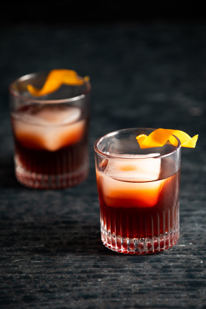 Two Espresso Negroni Cocktails in rocks glasses with an orange peel as garnish. Deep red in color. Espresso Negroni Cocktail Recipe
