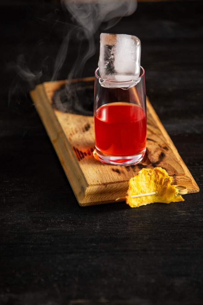 A cocktail red in color sits on a wooden board that is slightly burnt with smoke pouring out. Coo Coo for Coconuts Negroni