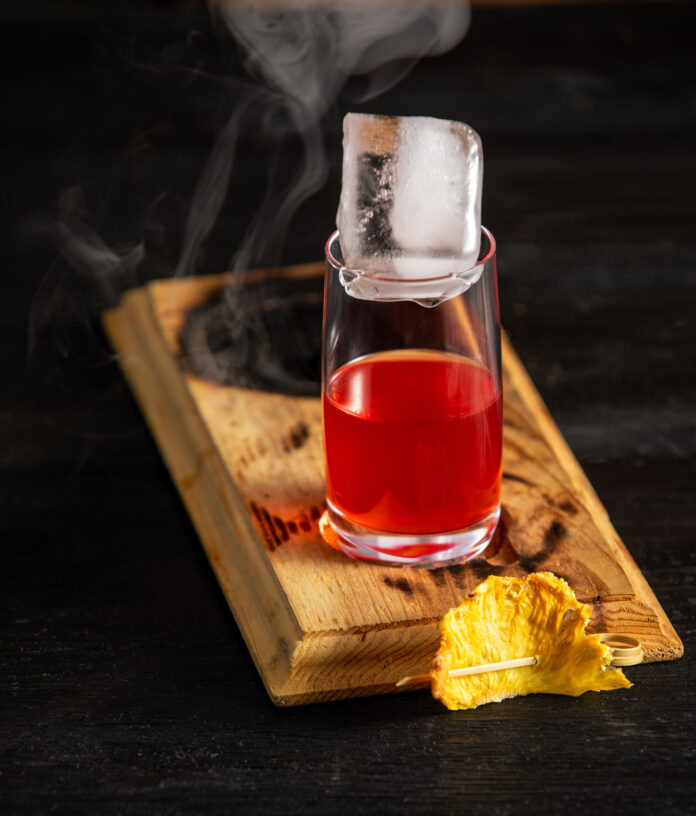 A cocktail red in color sits on a wooden board that is slightly burnt with smoke pouring out. Coo Coo for Coconuts Negroni