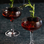 Two dark red Negroni cocktails in tall glasses garnished with rosemary and blackberry