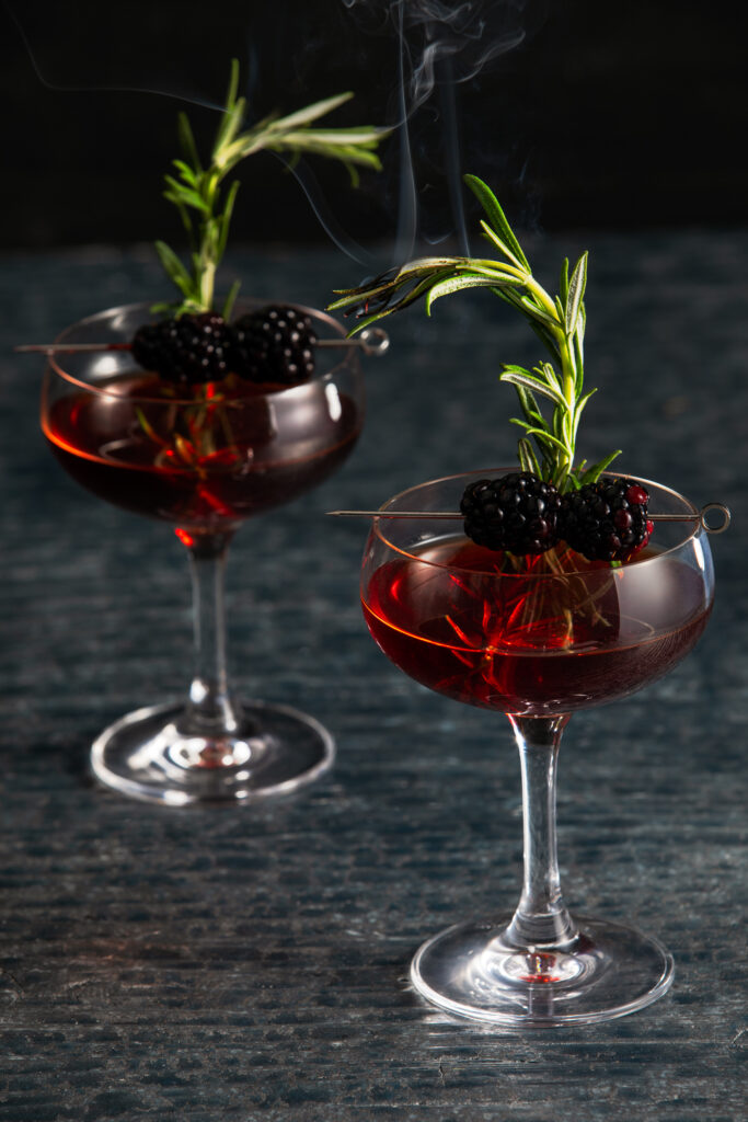 Two dark red Negroni cocktails in tall glasses garnished with rosemary and blackberry