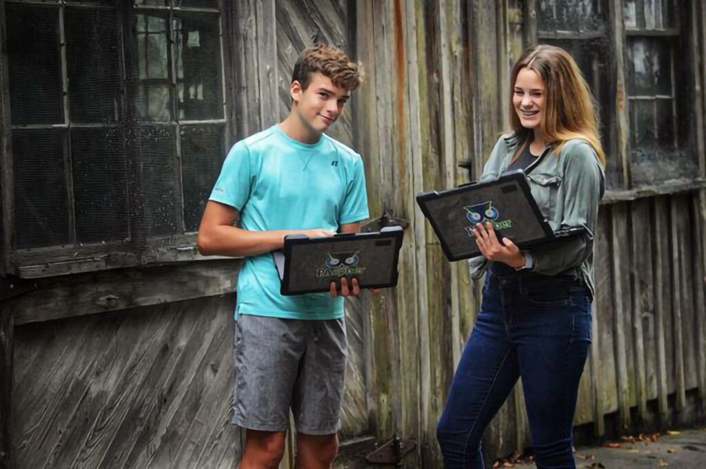 Two older children standing infirm of a wooden structure holding laptop computers with PA Cyber stickers on them. Pittsburgh schools culture