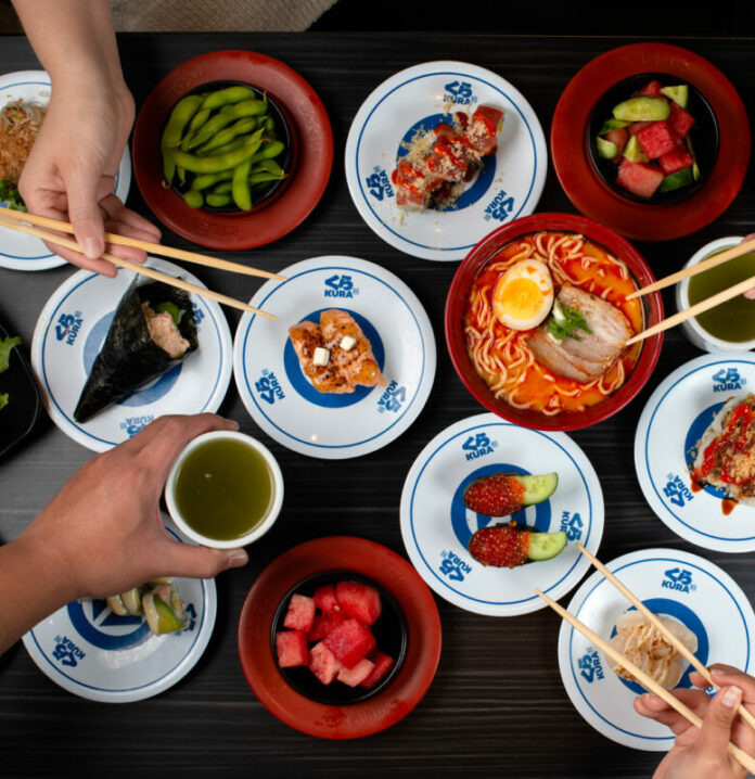 Four hands, three with chopsticks, hover over a table full of Japanese cuisine