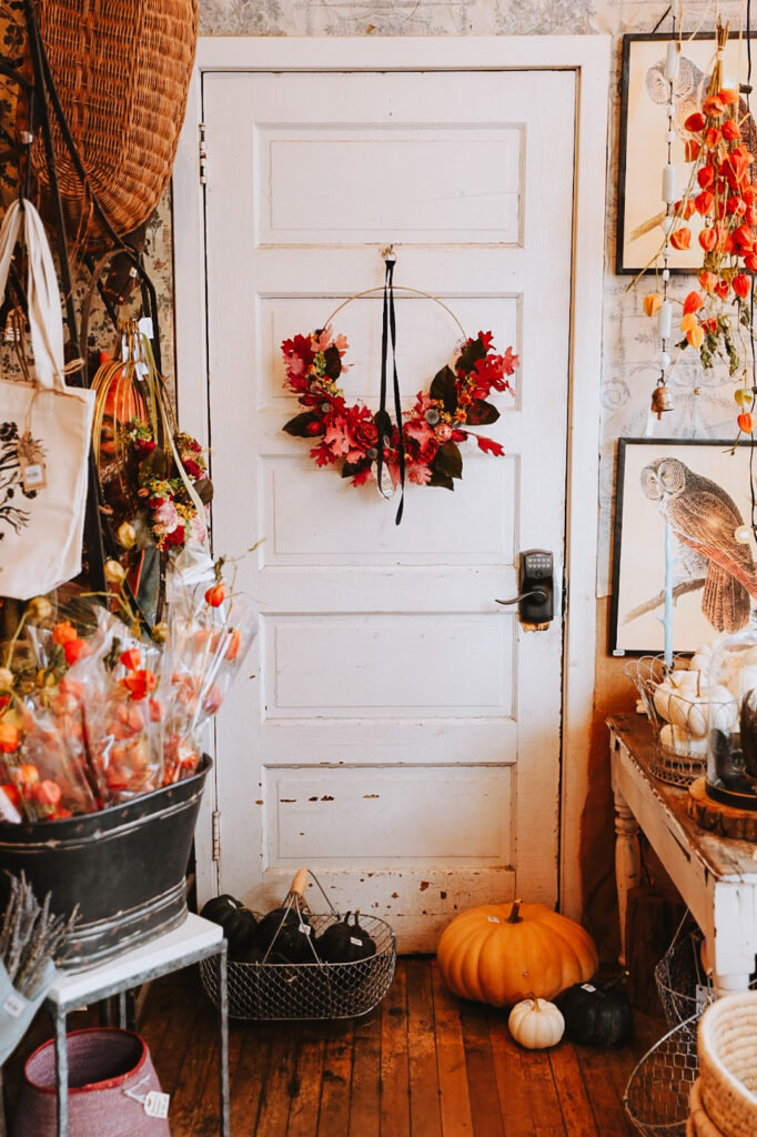 A white door with a leaf wreath is surrounded by fall decor like pumpkins