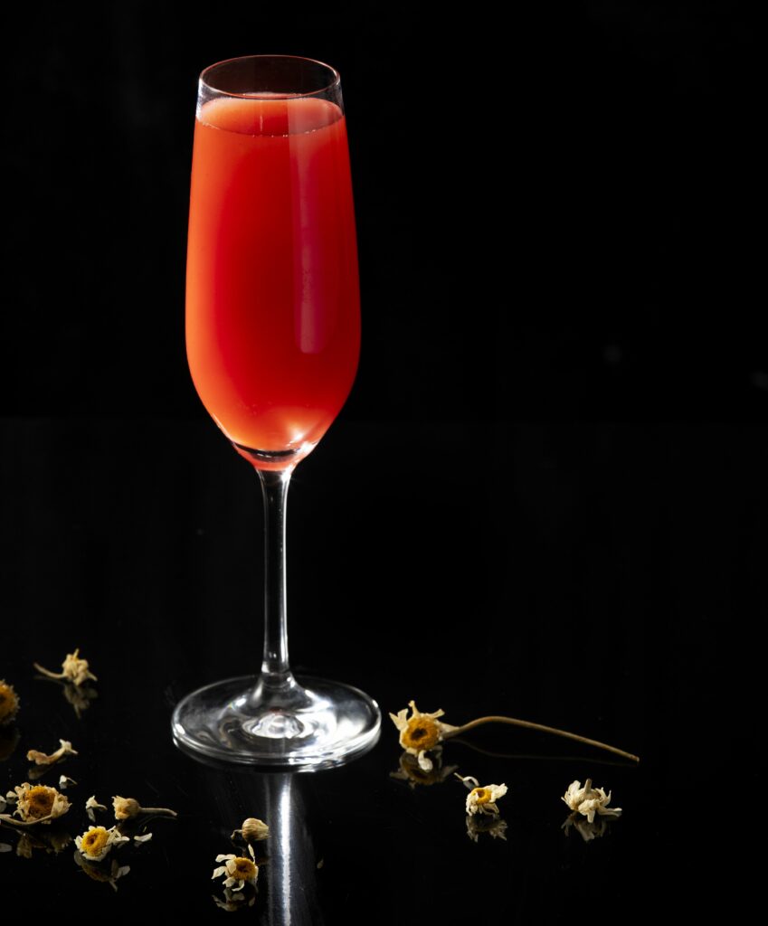 A deep pink, almost ruby red cocktail in a champagne flute on a black reflective surface surrounded by a handful of scattered dried chamomile flowers.