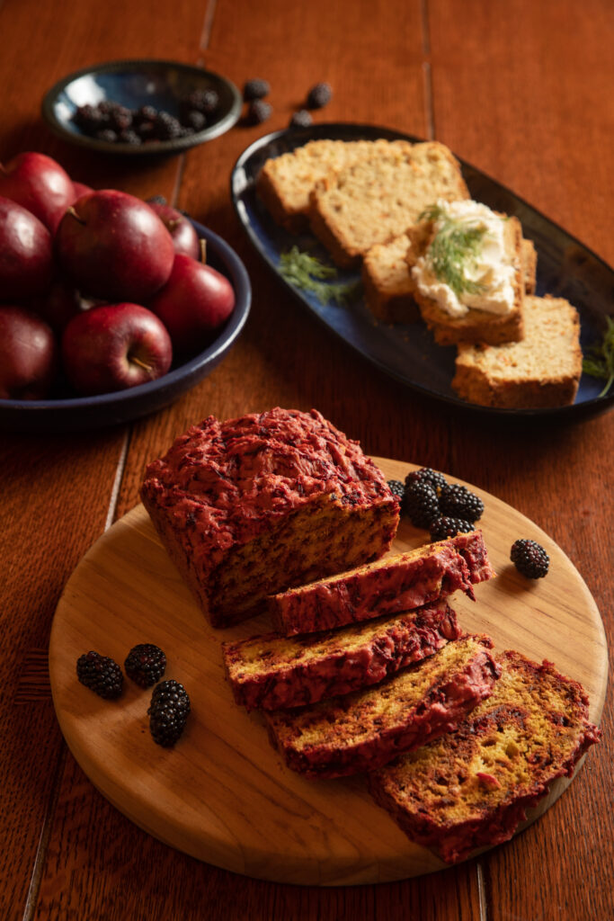 a loaf of bread sits cut up on a round wooden plate next to apples and blackberries. Kitchen Garden Quick Bread