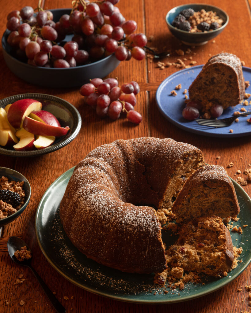 A bundt cake sits on a green plate with a third cut out. Various fruits and sits on plates around the cake. Granola Breakfast Cake Recipe
