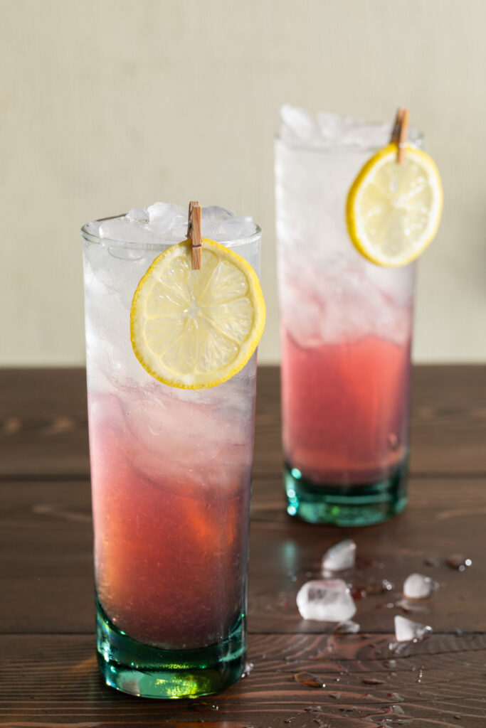 A wooden surface with 2 cocktails in green tinted collins glasses, garnished with a lemon wheel and a tiny clothespin.