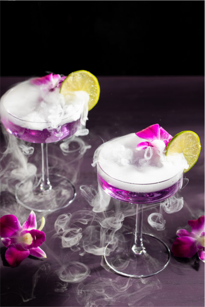 Two glasses with purple liquid inside are topped with little botanical pink flowers and lemon slices with smoke pouring overtop.
