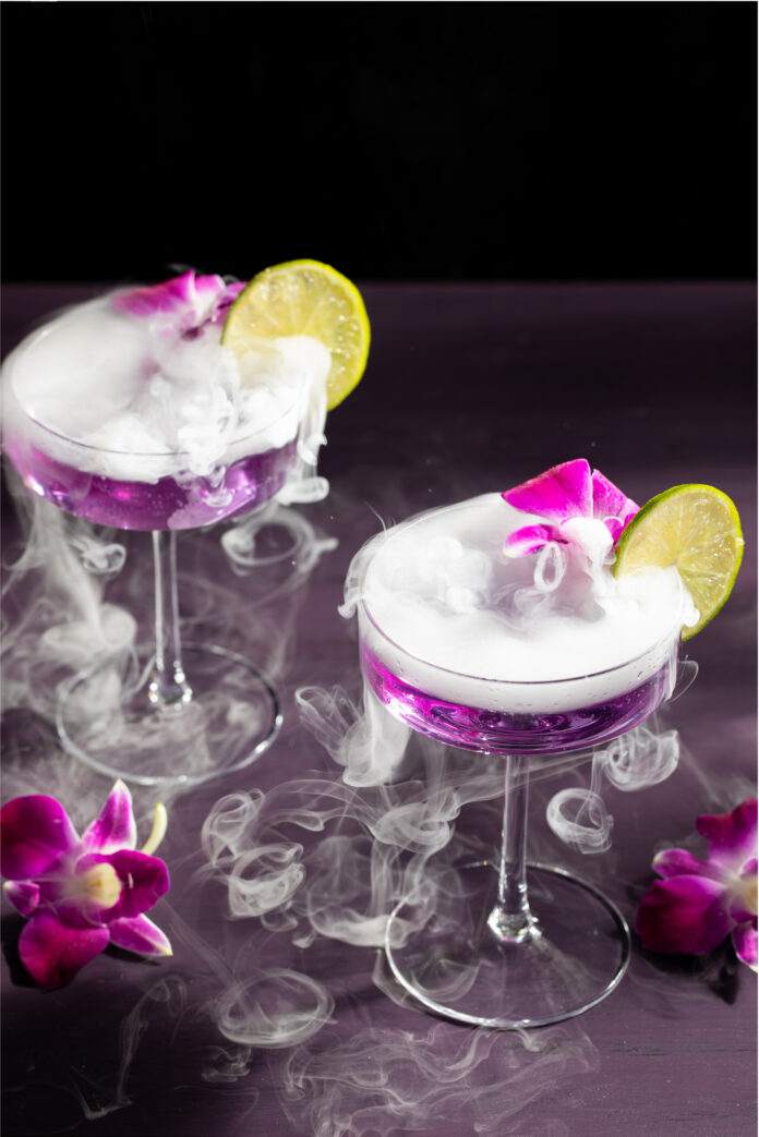 Two glasses with purple liquid inside are topped with little botanical pink flowers and lemon slices with smoke pouring over the cocktail.