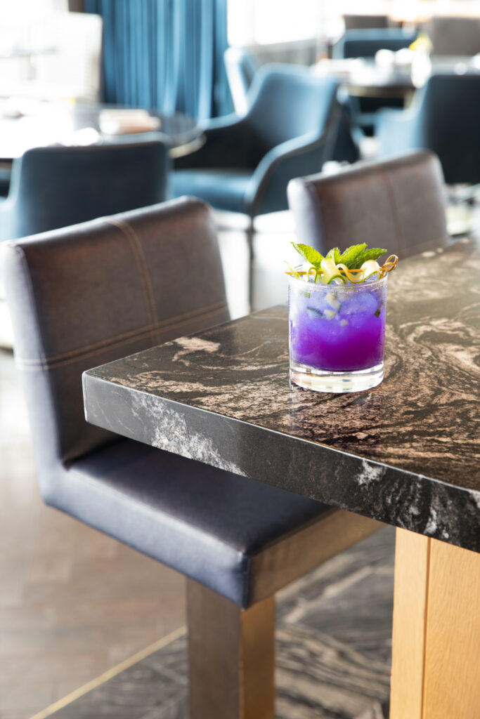 a cocktail, dark purple in color, sits on a black marble table. Wild Blossom Cocktail recipe from The Oaklander. pea flower cocktail