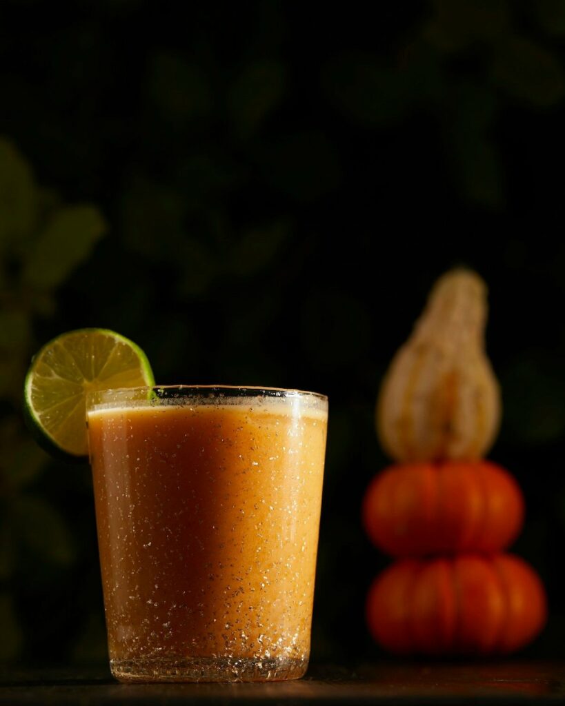 a cocktail orange in color sits in a rocks glass. two pumpkins and a squash are in the background. the cocktail is a pumpkin spice cocktail, a mai tai