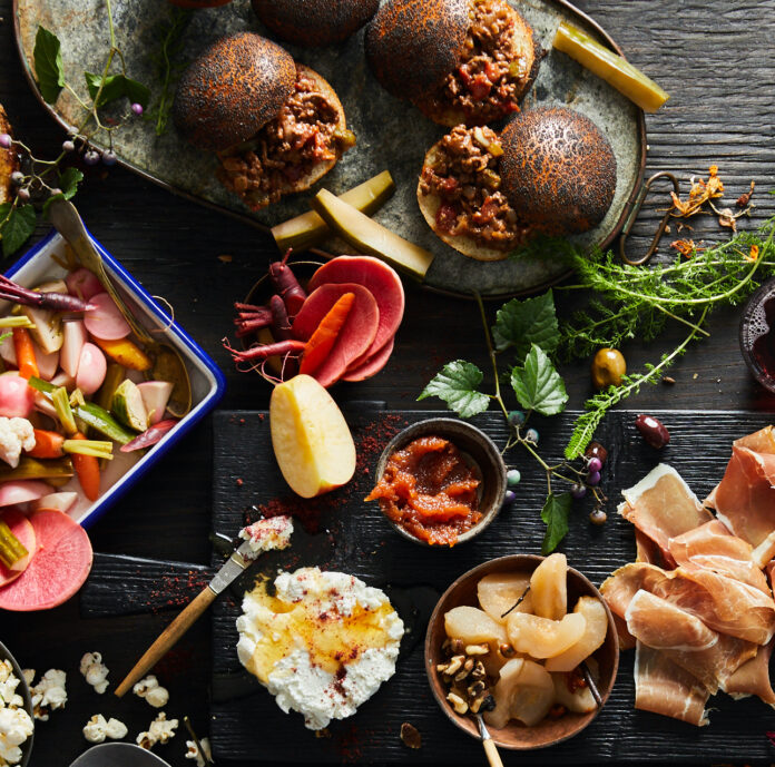 Venison Sloppies, or wild game sloppie joes on a green plate at the top corner of the frame, next to a spread of various other foods like fresh fruits and veggies