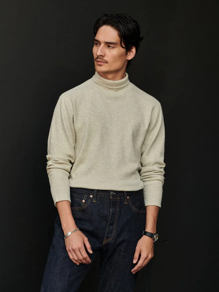 A dark-haired man models a tan turtleneck styled with dark pants.