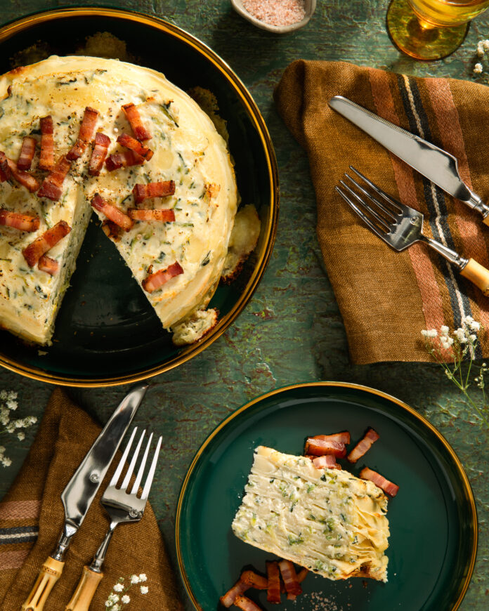 An aerial view of a Savory Zucchini Crepe Cake on a black plate with a slice cut out of it. The slice sits on a smaller green plate to the right.