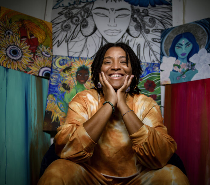 A young black woman in a tie dye outfit sits with smiling, center of the frame, with her hands on her face and her elbows on her knees.