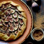 A galette with sliced figs on a terra cotta colored plate with figs and spice on a dark surface