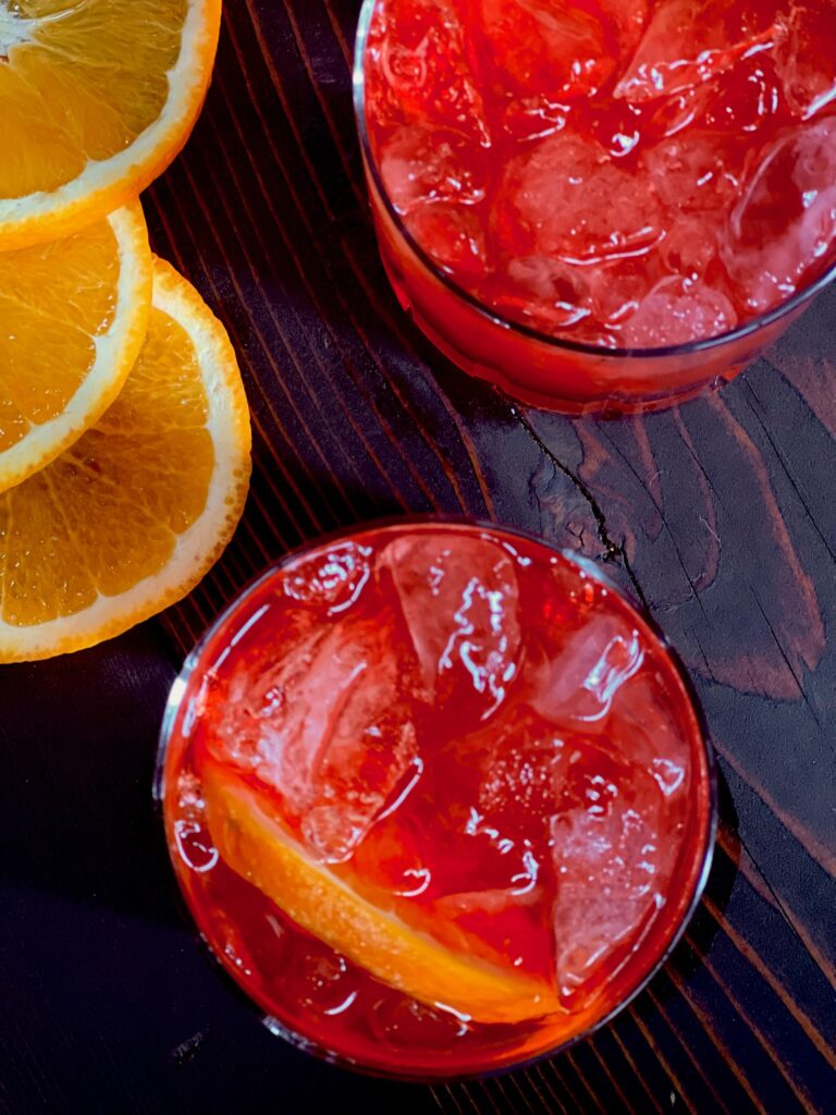 A close down shot of two red cocktails garnished with orange and a fan of orange slices at the top left.