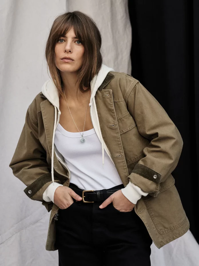 A brown-haired woman in a Buck Mason tan jacket with a hood and white t shirt.